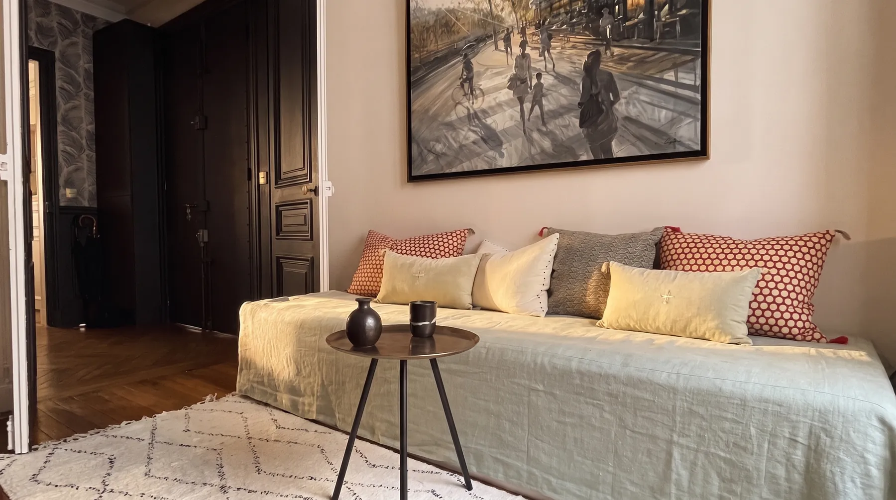 La pièce d'appoint vue en largeur : banquette habillée de lin vert d'eau, coussins dépareillés, grande toile encadrée, guéridon rond en laiton et tapis berbère