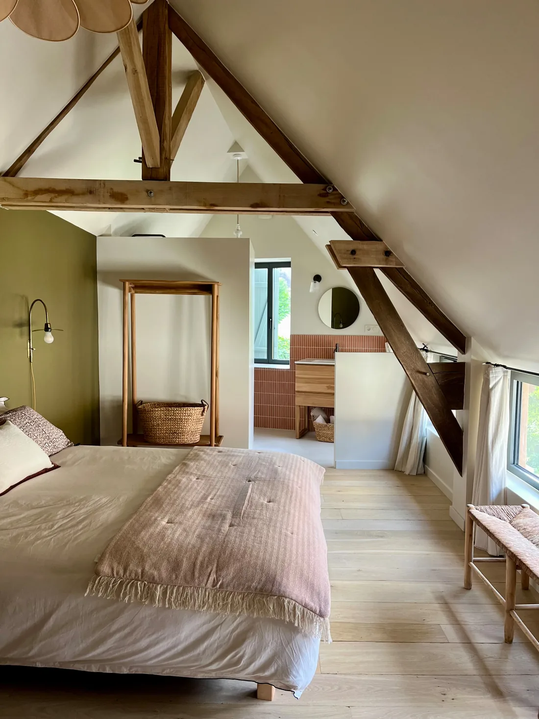 Chambre sous charpente : poutres et fermes apparentes, lit bas habillé de lin, cabine de douche dans l'angle et fenêtre donnant sur le jardin
