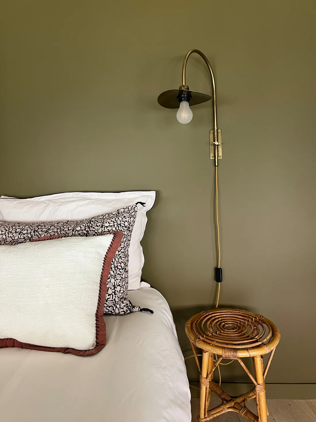 Détail de la chambre principale : mur kaki, liseuse de laiton en col de cygne, oreillers blancs à passepoil fleuri et tabouret de rotin en chevet