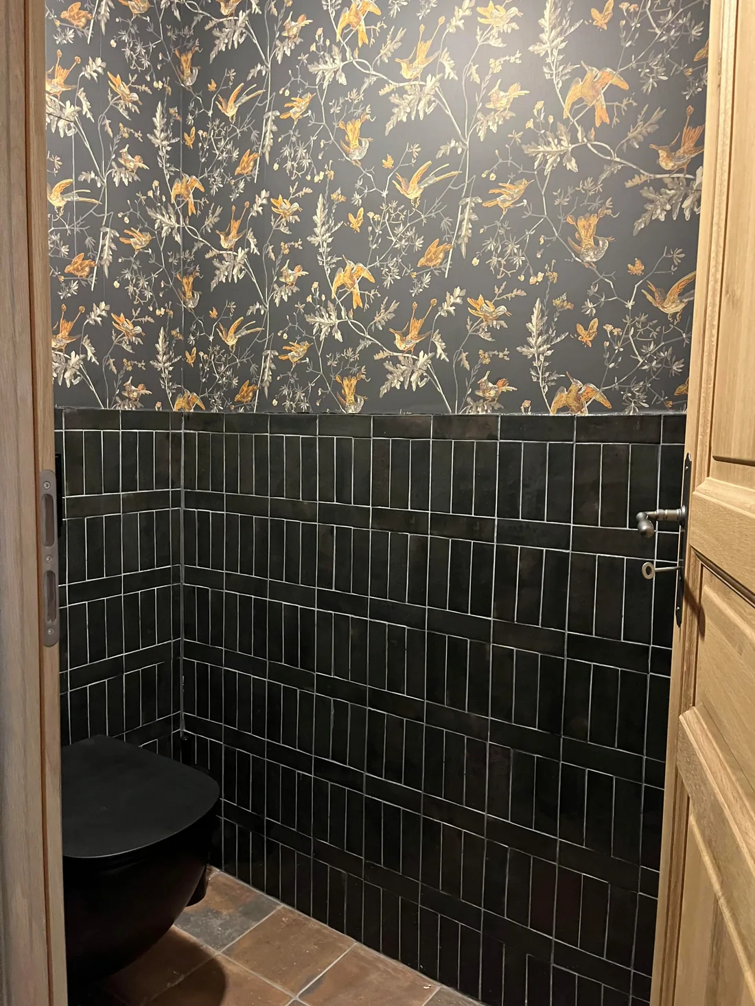 La douche en zellige noir brillant, receveur de tomettes anciennes, ouverte sur un mur de papier peint sombre à oiseaux et feuillages dorés