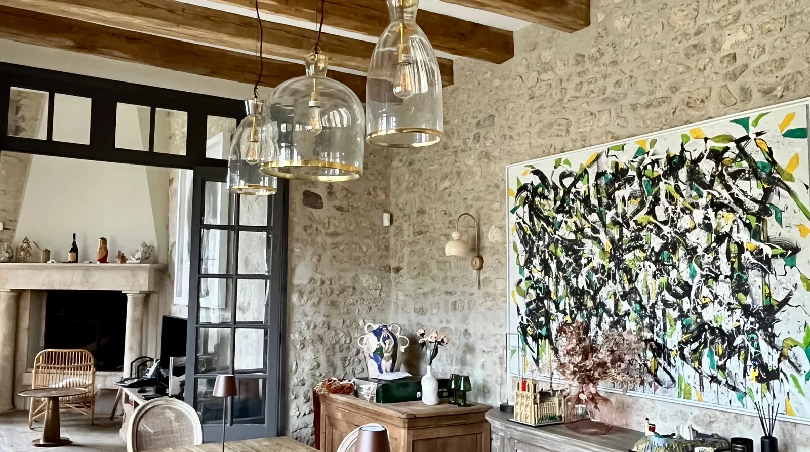 Le séjour de la maison : table de ferme en bois brut sous des poutres apparentes, mur de pierre, grande toile abstraite aux verts et jaunes, vaisselier ancien et suspensions de verre