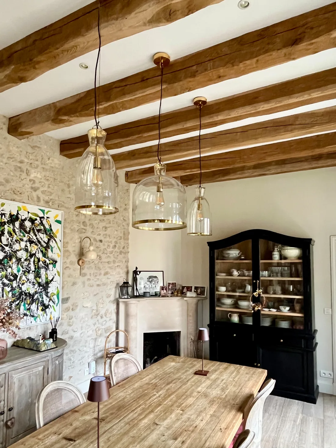La salle à manger : table de ferme en bois brut, poutres apparentes, suspensions de verre strié, cheminée blanche, grande toile abstraite et vaisselier noir garni de vaisselle ancienne