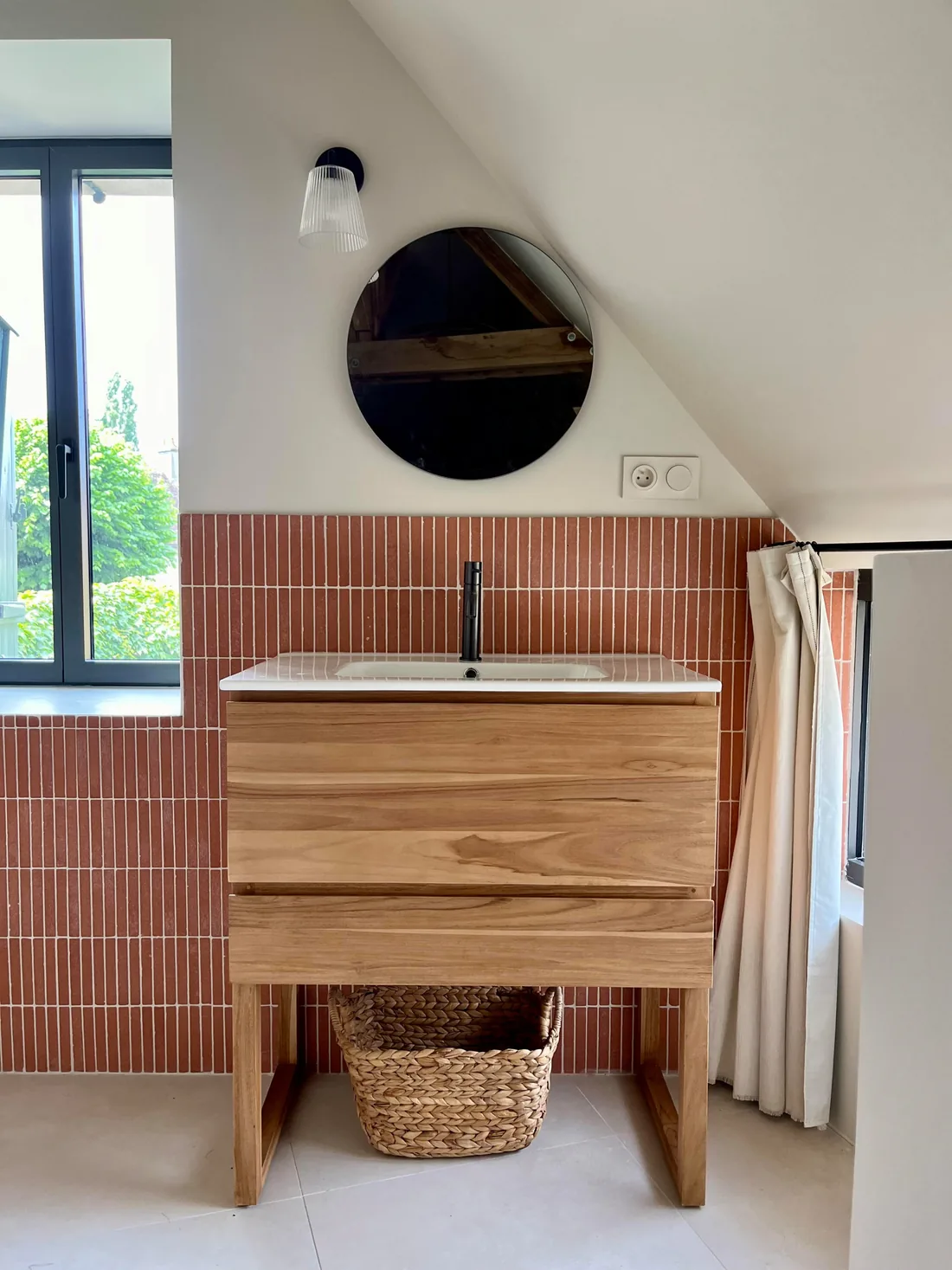 Salle de bains : meuble-vasque de bois brut à panier tressé, vasque blanche à robinet noir, mur de zellige terracotta posé en colonnes et rideau écru