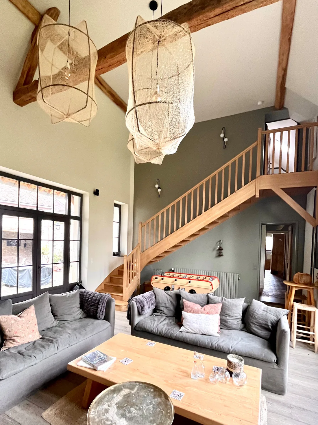 Le salon sous la mezzanine : canapés d'angle gris, verrière d'atelier en fond, escalier de bois vers la mezzanine et deux grandes suspensions de rotin ajouré