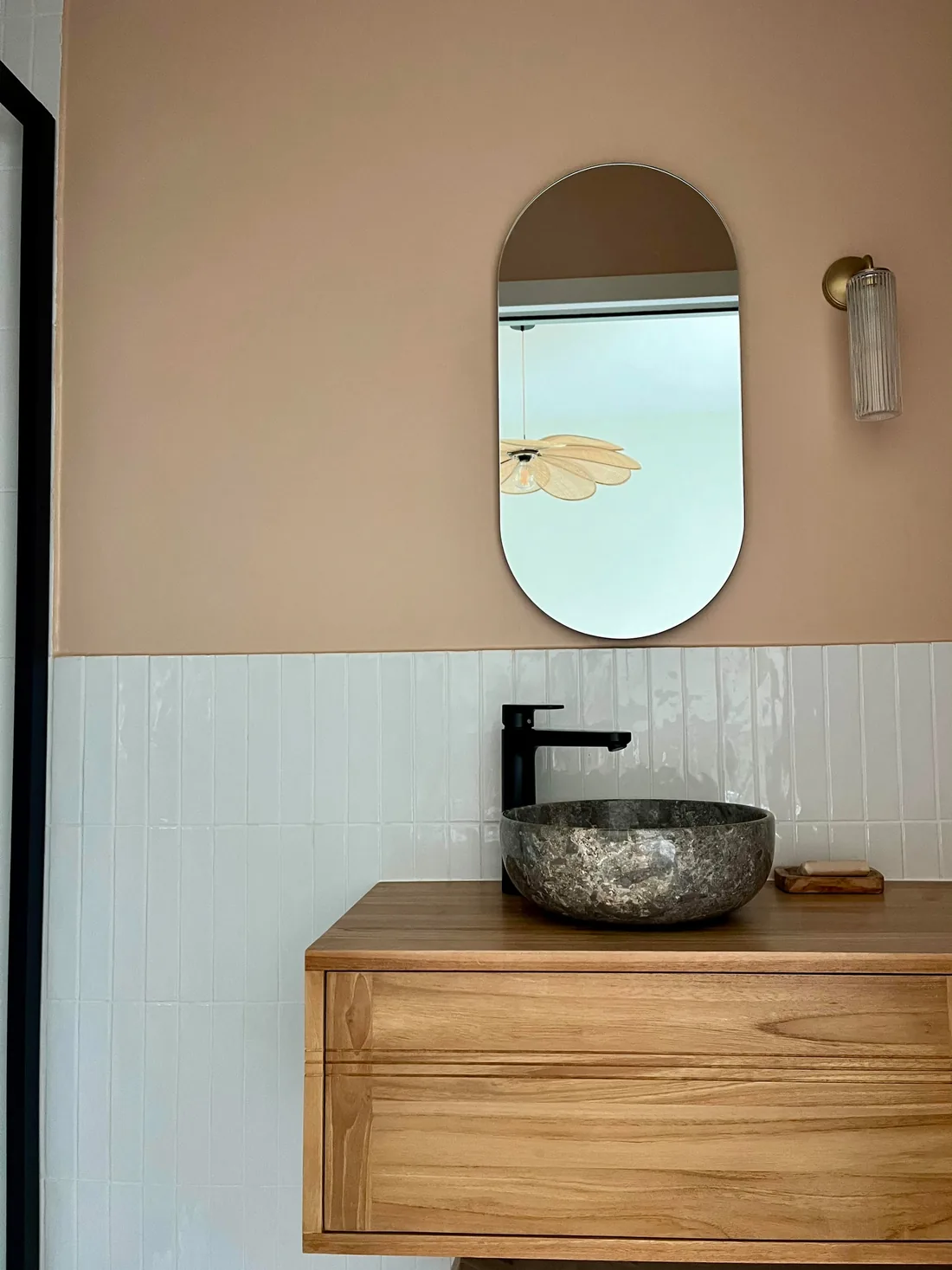 Salle d'eau : vasque de pierre grise taillée sur un meuble suspendu de chêne, miroir en arche sur un mur rose poudré, applique dorée et soubassement de carrelage blanc
