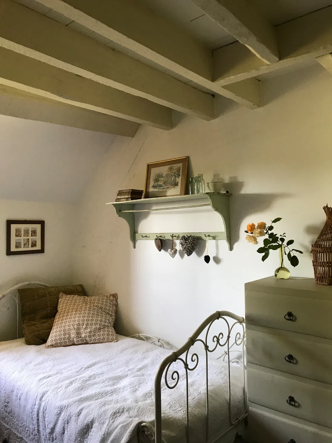 Chambre mansardée sous charpente : lit ancien en fer forgé blanc, linge de lit écru, étagère murale à crochets portant des fleurs séchées, commode de bois à trois tiroirs et gravures encadrées au mur