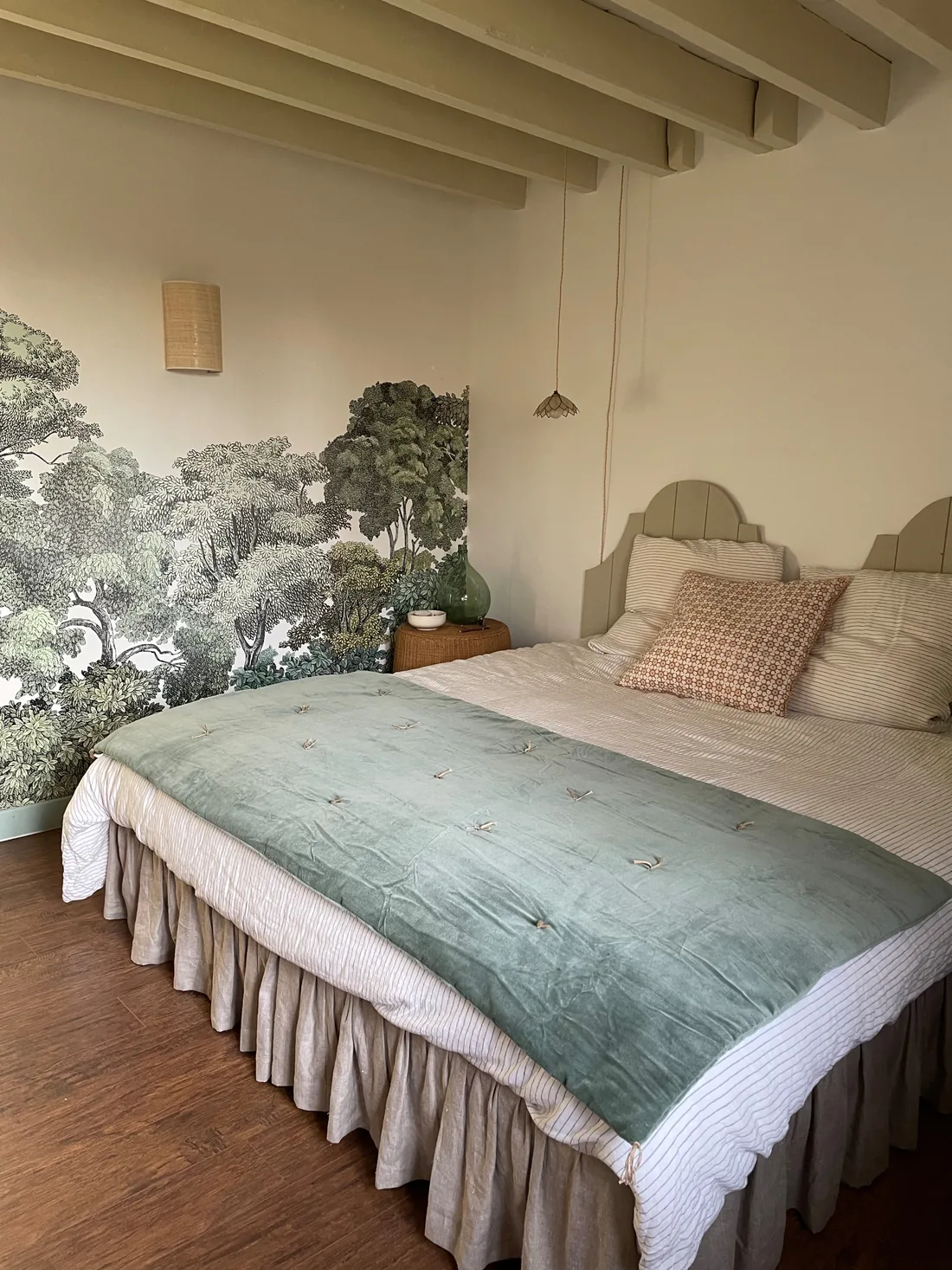 Chambre au papier peint panoramique représentant une forêt dessinée au trait noir sur fond clair, lit à jupe de lin blanc, couvre-lit de velours vert d'eau, coussins terracotta et poutres au plafond