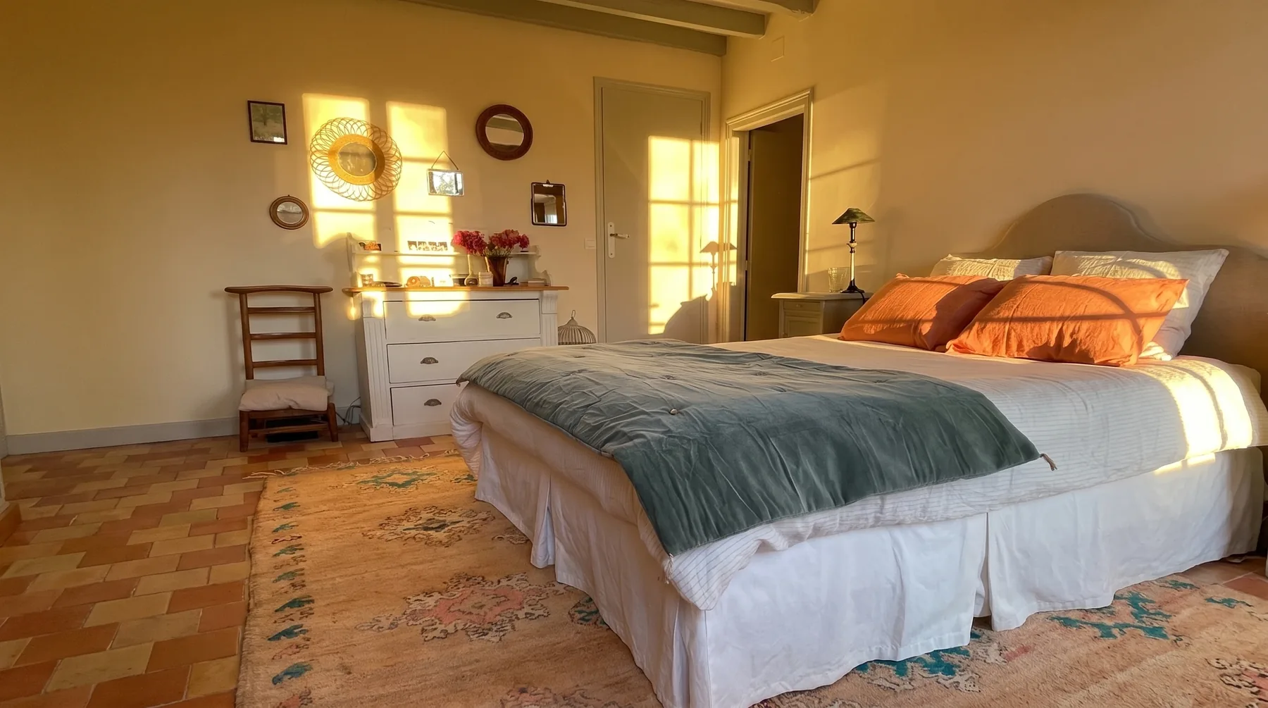 Chambre principale : tête de lit cintrée tendue de lin, oreillers orange et couvre-lit de velours vert d'eau, commode blanche ancienne sous des miroirs de rotin, tapis berbère aux tons fanés, sol en tomettes et lumière rasante de fin de journée