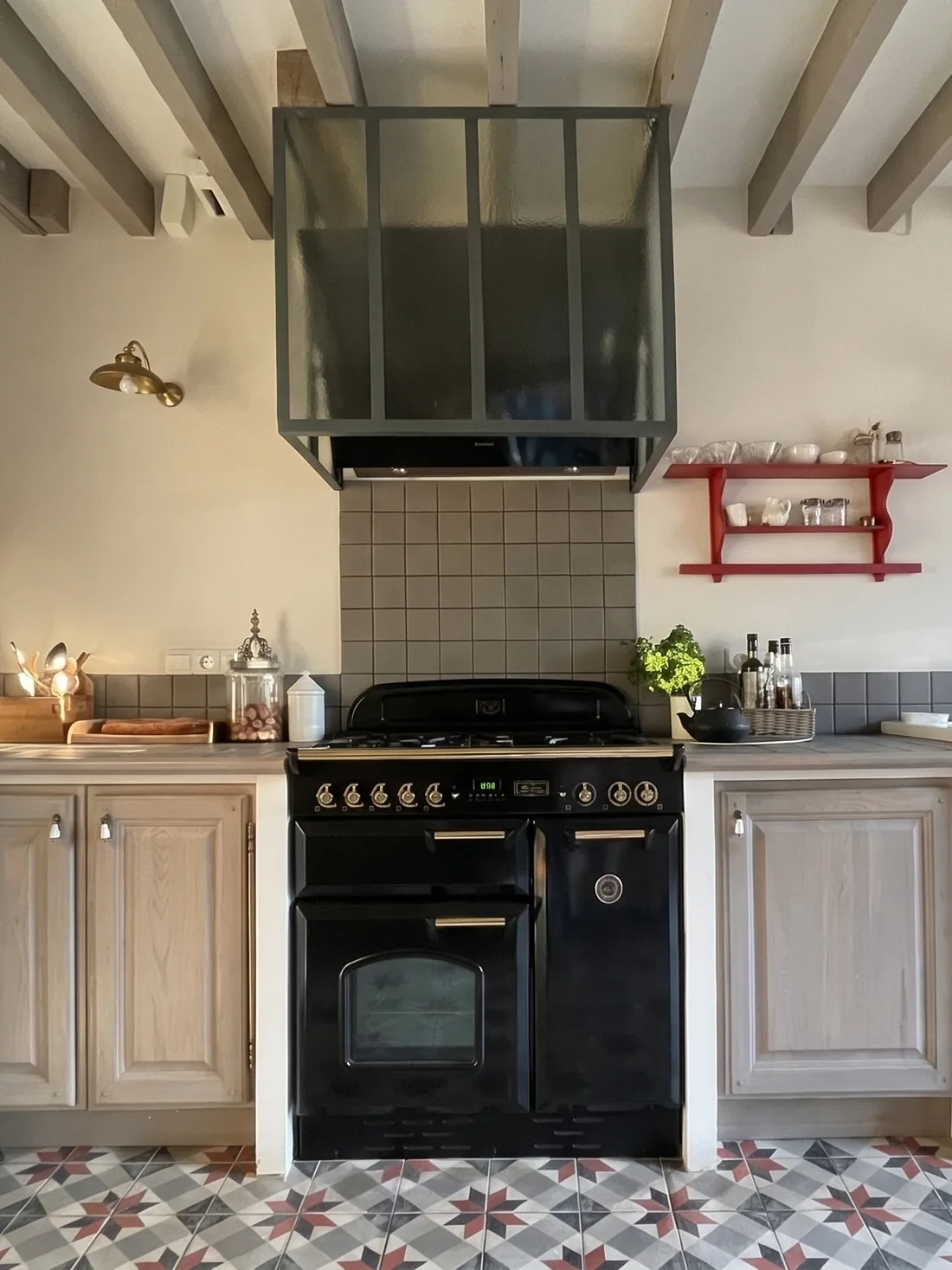 Le piano de cuisson noir à poignées de laiton sous sa hotte habillée d'une verrière vert sombre, crédence de carrelage gris mat, plan de travail de bois, étagères murales rouges garnies de vaisselle blanche