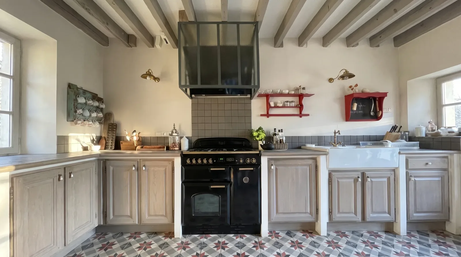 Cuisine de la maison de campagne : piano de cuisson noir sous une hotte habillée d'une verrière vert sombre, meubles de chêne cérusé, étagères murales rouges, appliques de laiton, plafond à poutres et sol en carreaux de ciment à motif d'étoiles