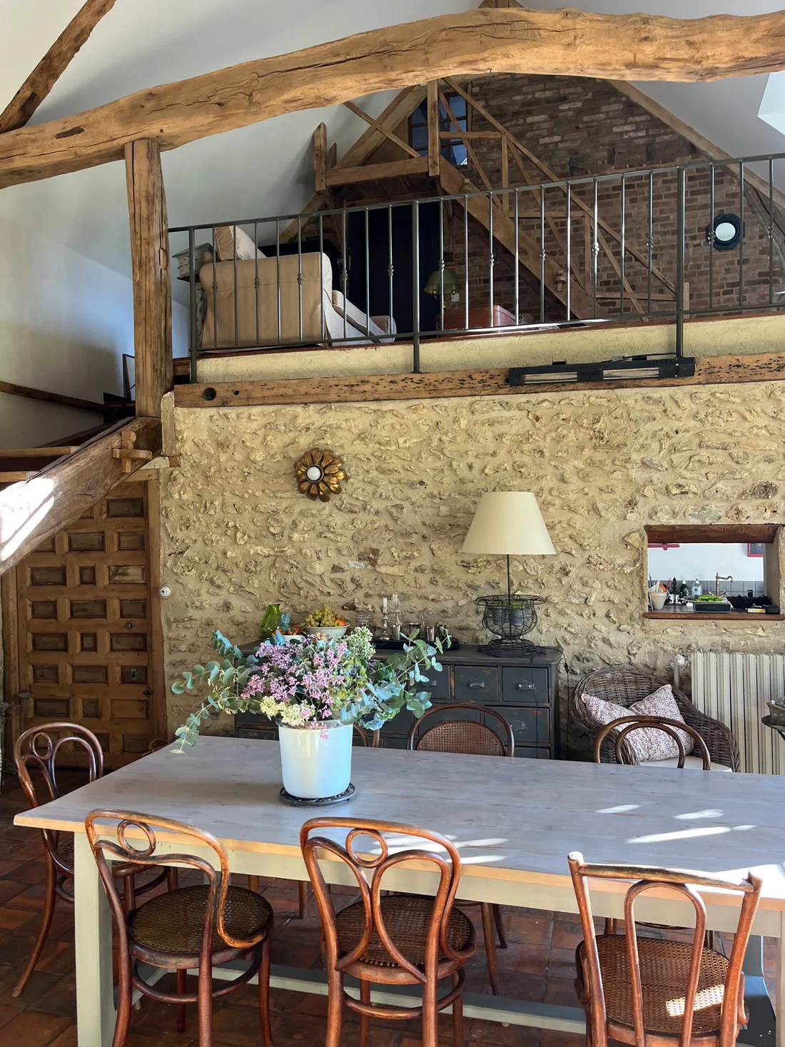 La salle à manger vue en hauteur : ferme de charpente ancienne, mezzanine à garde-corps de métal, mur de pierre, porte espagnole à caissons, commode patinée et table entourée de chaises bistrot