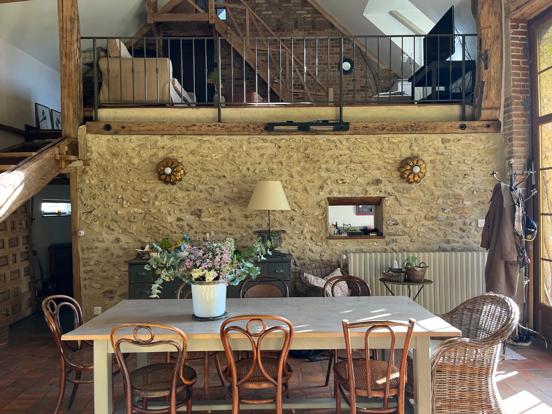 Salle à manger de la maison de campagne : mur de pierre apparente sur toute la hauteur, mezzanine à garde-corps de métal noir, grande table entourée de chaises bistrot dépareillées et de fauteuils de rotin, bouquet de fleurs des champs au centre