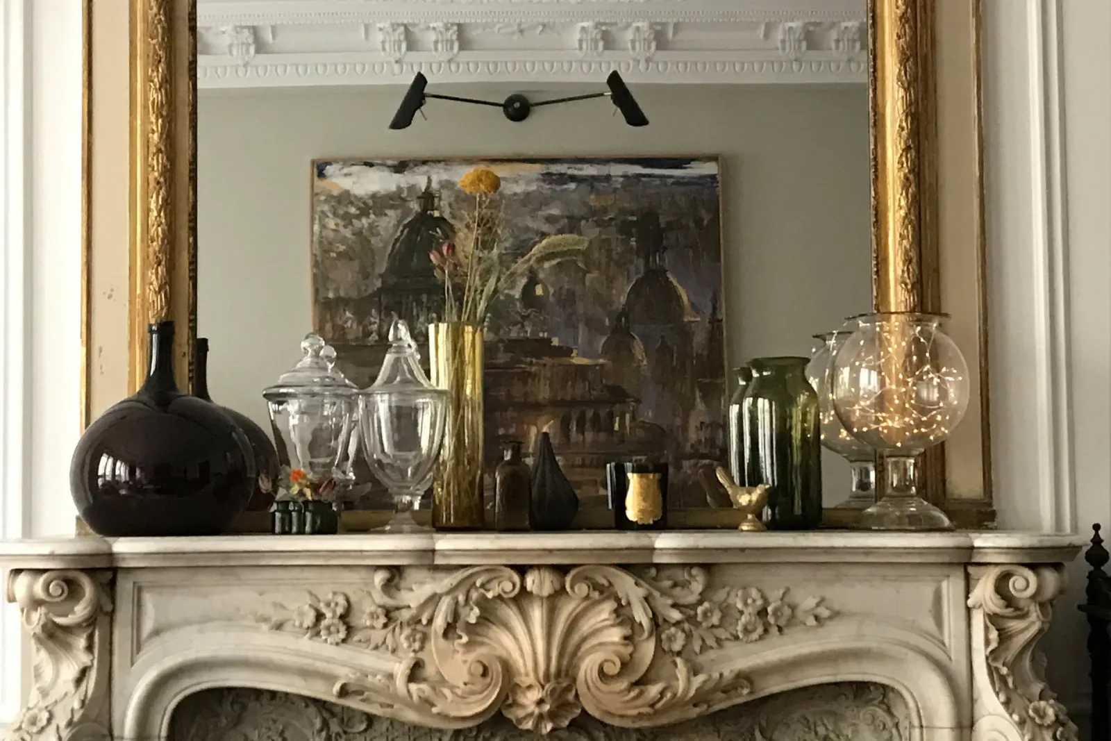Cheminée de marbre blanc sculptée d'une coquille dans un appartement haussmannien parisien, surmontée d'un trumeau doré et d'une collection de verreries anciennes