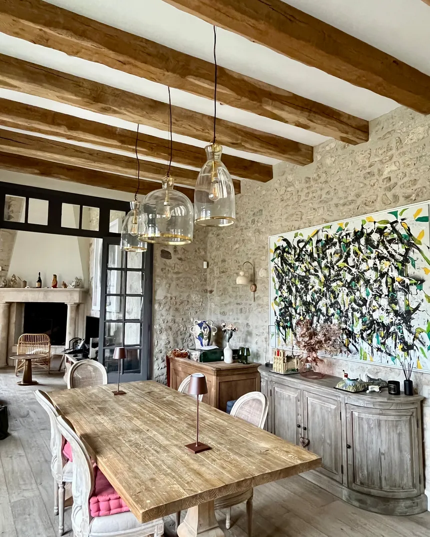 Salle à manger d'une maison de campagne près de Fontainebleau : mur en pierre apparente, poutres, grande table en bois brut et suspensions en verre