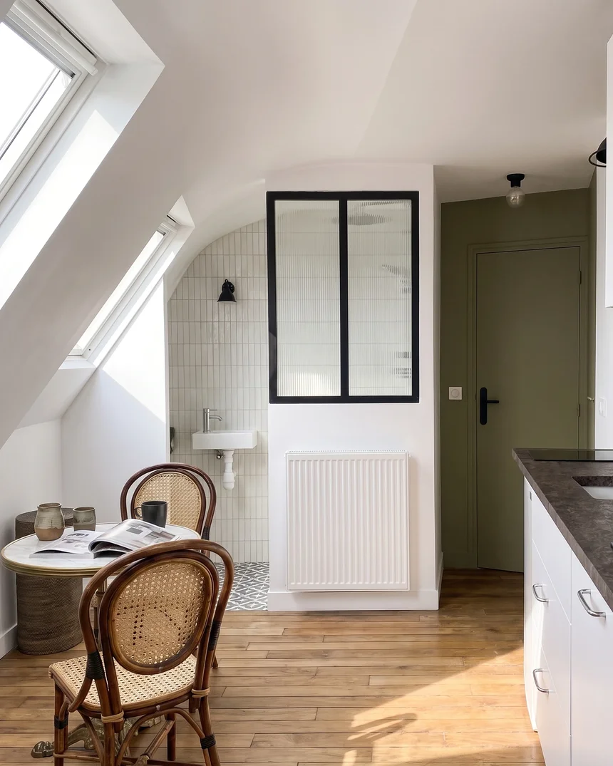 Vue en trois dimensions du studio de 10 m² sous les toits : cuisine linéaire blanche à plan de travail sombre, lit encastré sous la pente et tête de lit capitonnée