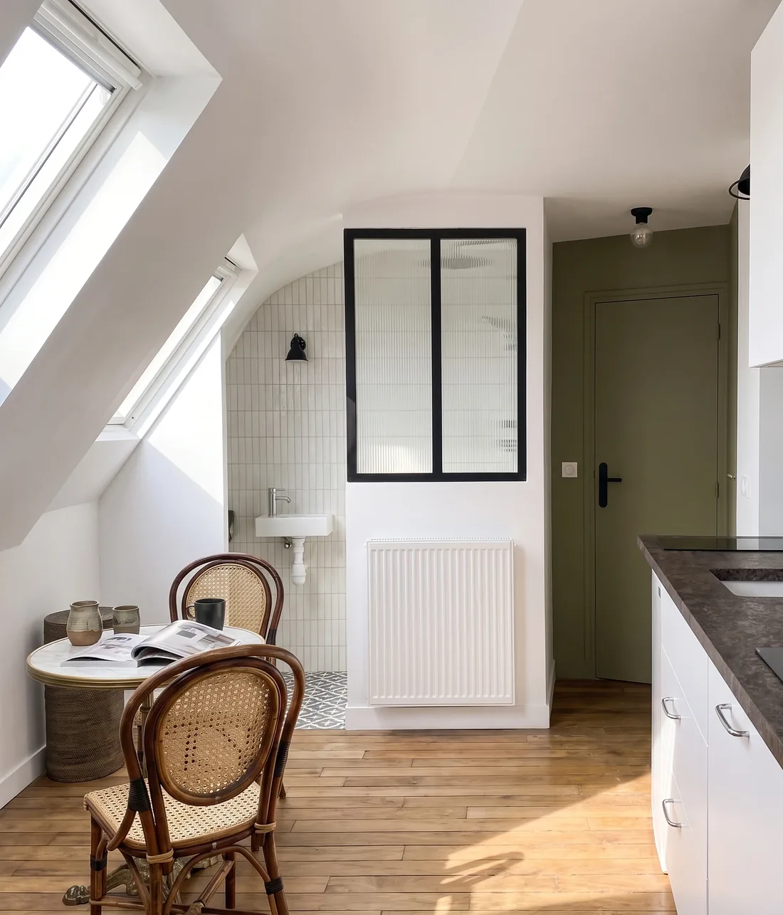 Vue du projet : la salle d'eau derrière sa verrière à châssis noir, lavabo et miroir éclairés, radiateur sous la cloison et kitchenette à plan sombre au premier plan