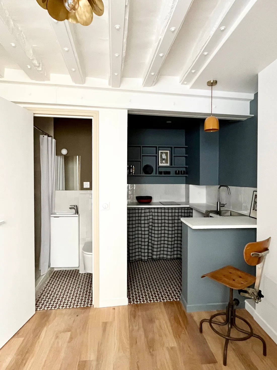 La cuisine : meubles bleu paon, plan de travail blanc, damier noir et blanc au sol, tabouret de bar en bois et métal et porte de la salle d'eau ouverte au fond