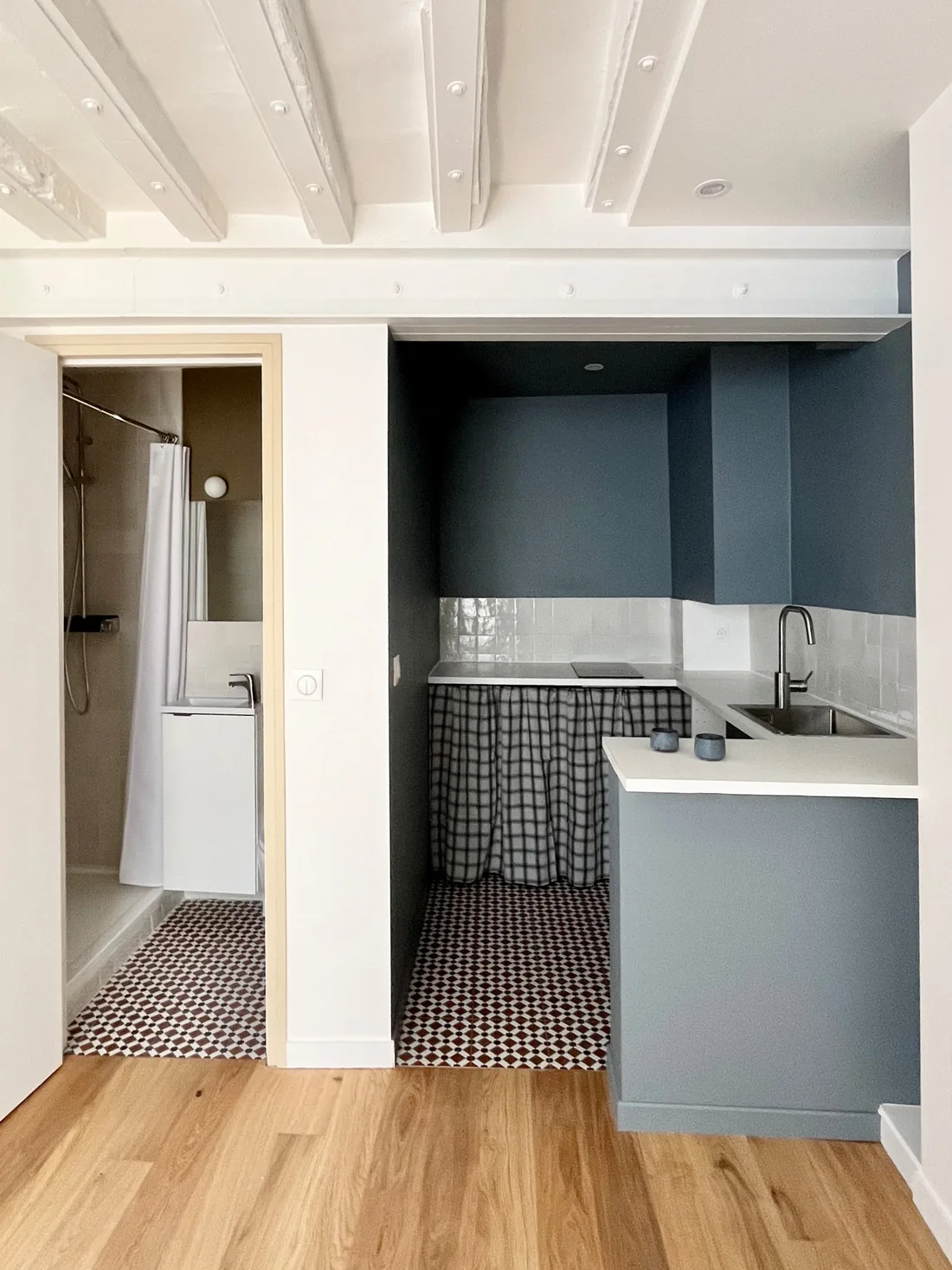 Détail de la cuisine : rideau à carreaux vichy gris sous l'évier, crédence de marbre clair, robinet noir et étagère bleu paon