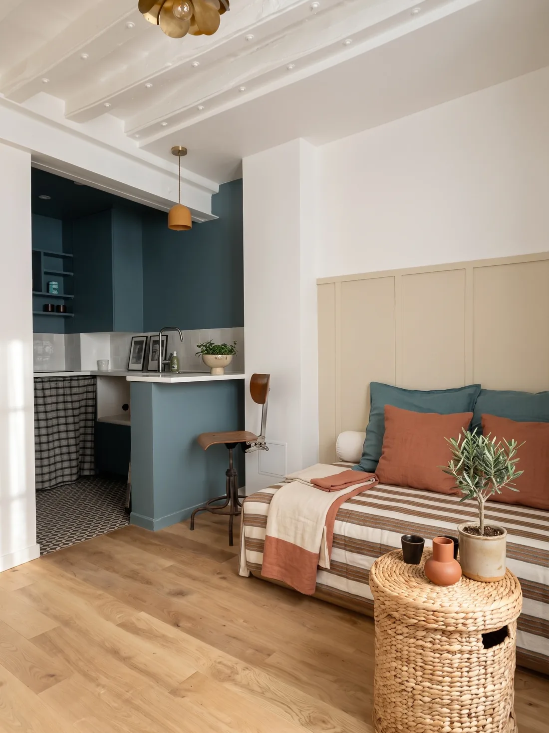 La cuisine bleu paon derrière son muret : tabouret d'atelier en bois et métal, rideau à carreaux sous l'évier, sol à motifs, suspension ocre, et au premier plan le lit rayé avec sa table d'appoint en fibre tressée et son olivier en pot