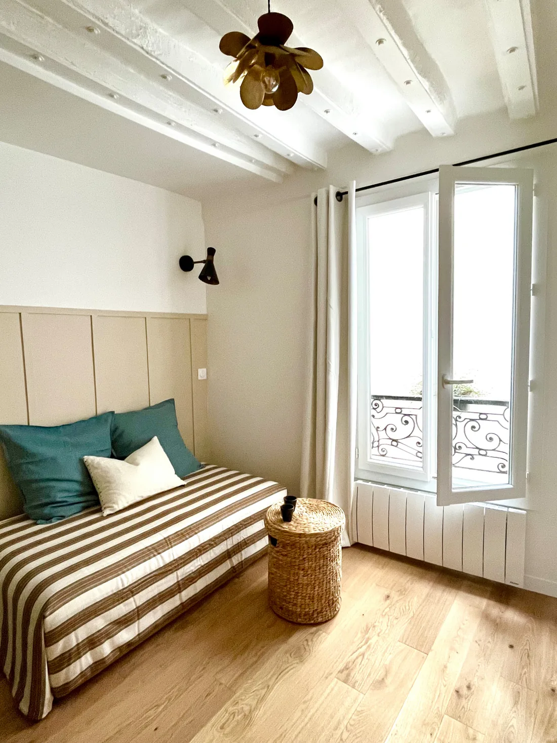 L'espace de vie : lit habillé d'un tissu rayé taupe et blanc avec coussins verts, lambris peint à mi-hauteur, applique noire, panier tressé et poutres blanches au plafond