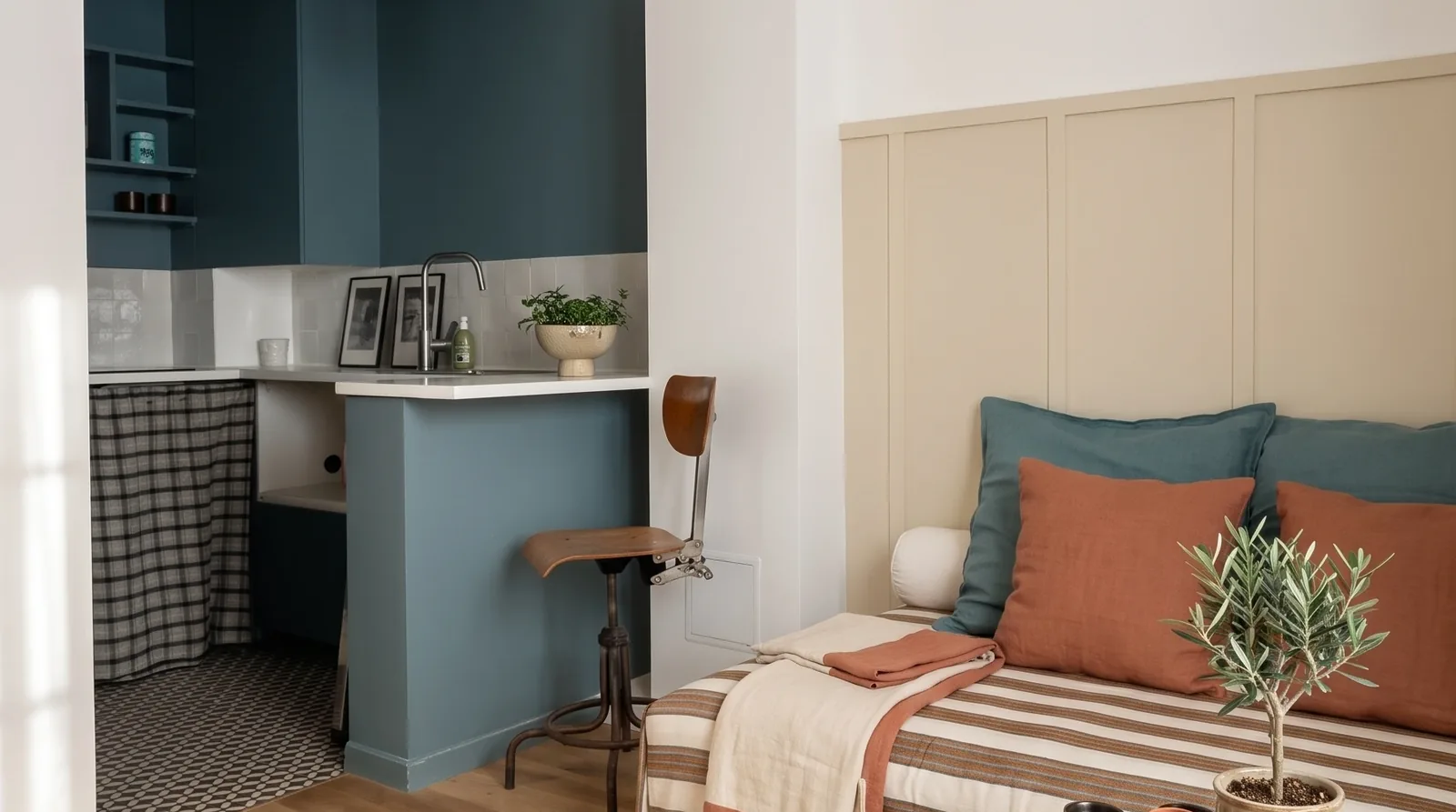 Le studio de 13 m² : la cuisine bleu paon derrière son muret avec tabouret d'atelier et suspension ocre, et le lit habillé d'un tissu rayé aux coussins bleu canard et terracotta, sous les poutres blanches