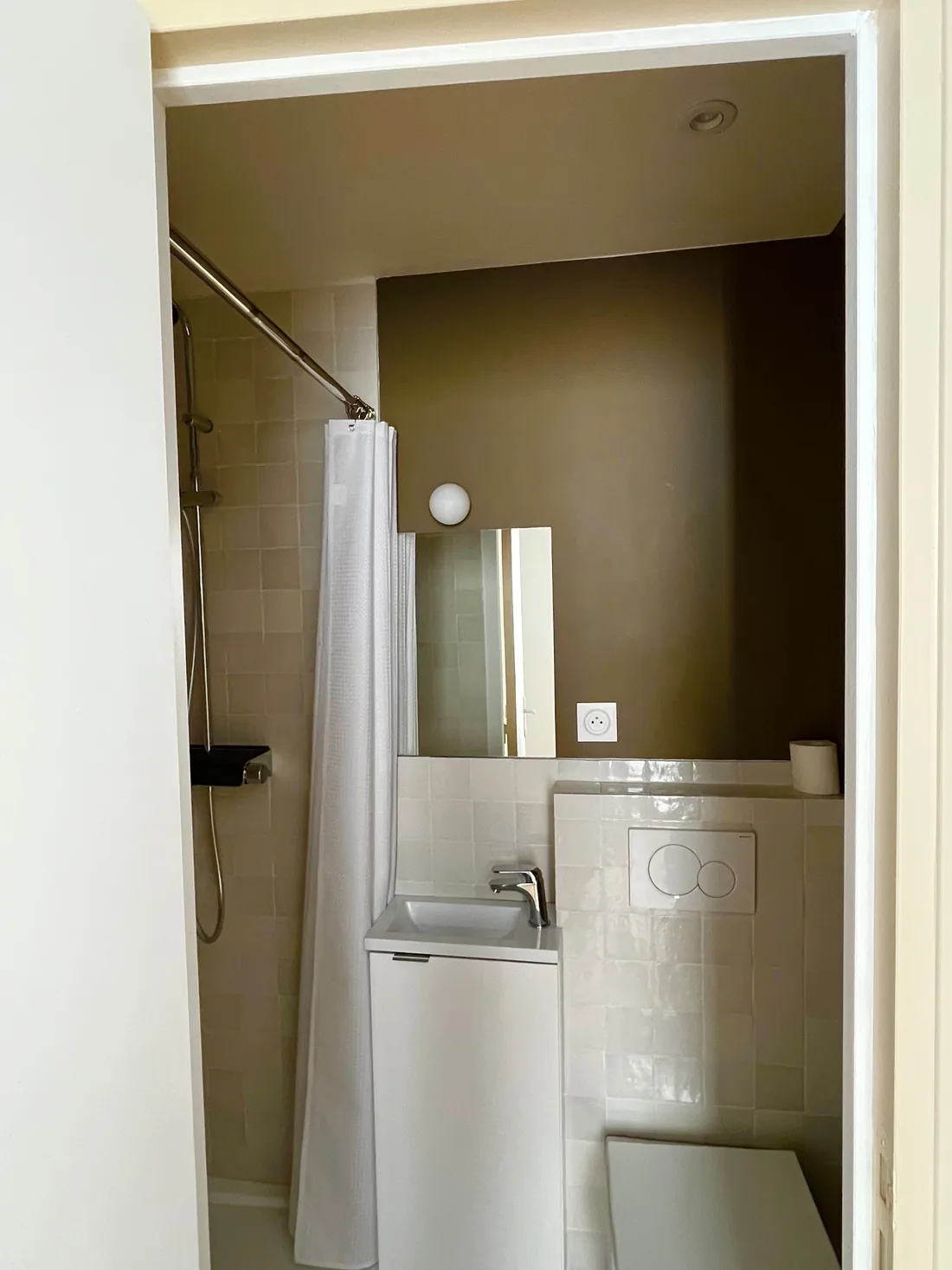 La salle d'eau de moins de 2 m² : douche à rideau blanc, lavabo d'angle avec miroir, murs kaki et porte coulissée