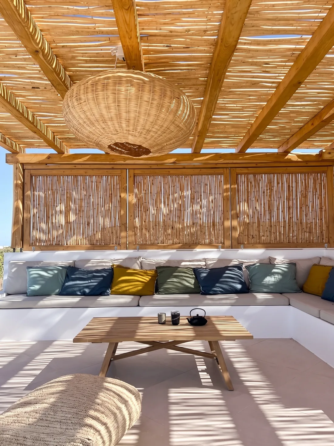 La banquette de la terrasse : coussins vert sapin, jaunes et bleus, table basse de bois brut, claustra de roseaux filtrant la lumière rasante et suspension en osier