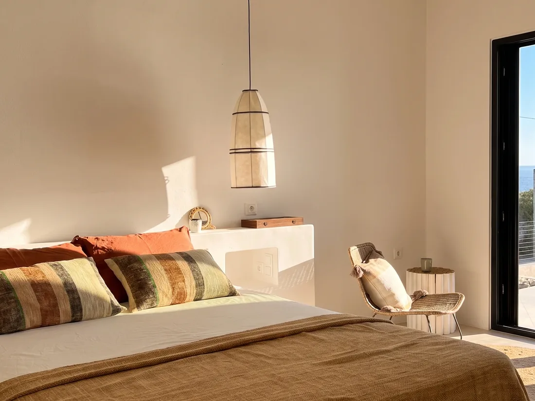 Chambre en fin de journée : lit habillé de lin et de jeté à carreaux, tête de lit maçonnée, suspension de paille tressée et lumière dorée du soir