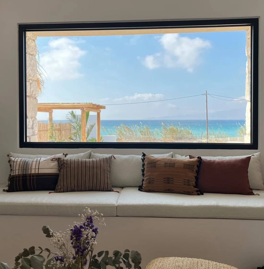 Une fenêtre à châssis noir cadrant la mer Égée, au-dessus d'une banquette maçonnée garnie de coussins rayés terracotta et écrus