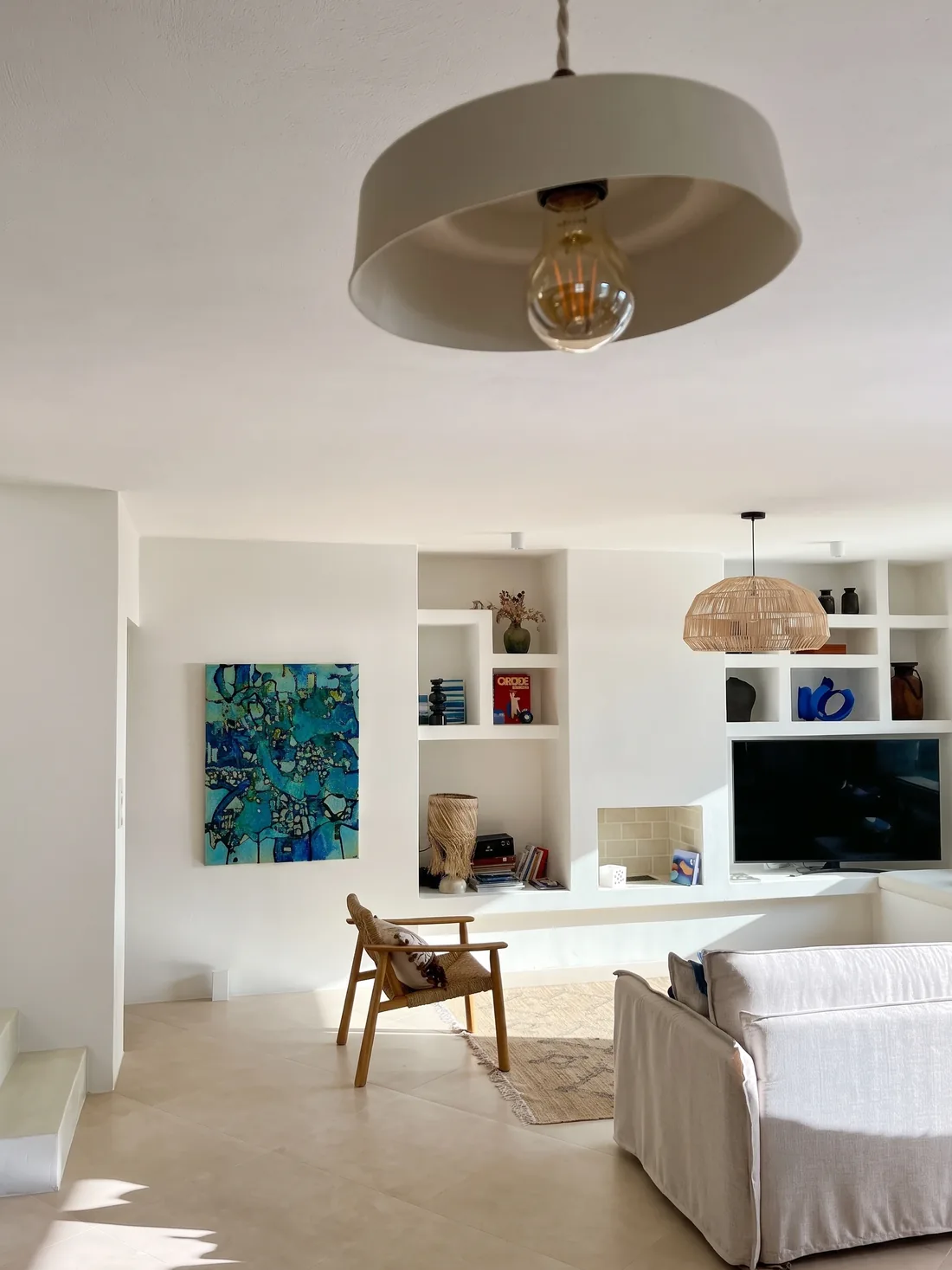 Le séjour blanc : banquettes maçonnées à coussins écrus, niches murales garnies d'objets, fauteuil de bois et toile aux bleus vifs accrochée au mur